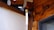 A wooden wall with visible beams and a hanging wind chime. The chime features a brass sphere and a vertical, flat white piece. The setting is in a tranquil corner with natural wood textures accentuated with soft light.