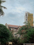A two-story house with a red-tiled roof is partially obscured by lush green trees. In the background, a tall building is under construction, wrapped in yellow scaffolding netting. A palm tree is visible in the left foreground, and the sky above is cloudy and overcast.