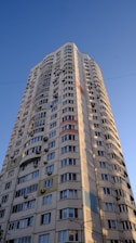 A tall residential building with numerous windows and air conditioning units attached to the exterior walls. The building features a modern architectural design with curved edges and colorful panels integrated into the facade. It stands against a clear blue sky, suggesting a sunny day.