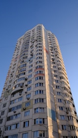 A tall residential building with numerous windows and air conditioning units attached to the exterior walls. The building features a modern architectural design with curved edges and colorful panels integrated into the facade. It stands against a clear blue sky, suggesting a sunny day.