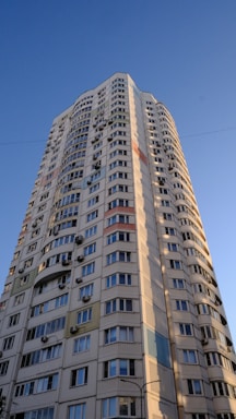A tall residential building with numerous windows and air conditioning units attached to the exterior walls. The building features a modern architectural design with curved edges and colorful panels integrated into the facade. It stands against a clear blue sky, suggesting a sunny day.
