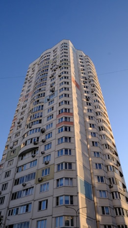 A tall residential building with numerous windows and air conditioning units attached to the exterior walls. The building features a modern architectural design with curved edges and colorful panels integrated into the facade. It stands against a clear blue sky, suggesting a sunny day.