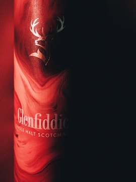 A strikingly lit Glenfiddich Single Malt Scotch Whisky bottle is partially shown, with a red and black swirling pattern. The iconic stag logo is prominently displayed in a metallic finish, giving it an elegant look.