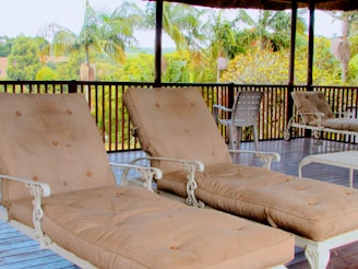 A veranda featuring two cushioned lounge chairs with metal frames positioned on a wooden deck. In the background, lush palm trees and various green foliage are visible, providing a tropical atmosphere. Additional seating can be seen, including plastic chairs and another lounge chair.
