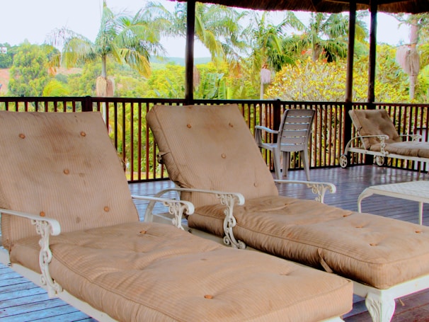 A veranda featuring two cushioned lounge chairs with metal frames positioned on a wooden deck. In the background, lush palm trees and various green foliage are visible, providing a tropical atmosphere. Additional seating can be seen, including plastic chairs and another lounge chair.