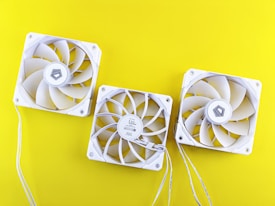 Three white computer cooling fans are arranged on a bright yellow background. Each fan has a similar design with multiple blades and a square frame. Thin white wires extend from the fans.