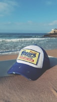 A blue and white baseball cap with the words 'Western New York Vintage' and 'Adventures Begin' written on a rectangular patch is placed on a sandy beach. In the background, gentle ocean waves roll onto the shore under a clear blue sky. A rocky outcrop can be seen jutting into the ocean from the right.