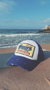 A blue and white baseball cap with the words 'Western New York Vintage' and 'Adventures Begin' written on a rectangular patch is placed on a sandy beach. In the background, gentle ocean waves roll onto the shore under a clear blue sky. A rocky outcrop can be seen jutting into the ocean from the right.
