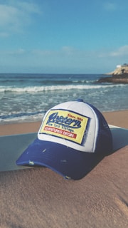 A blue and white baseball cap with the words 'Western New York Vintage' and 'Adventures Begin' written on a rectangular patch is placed on a sandy beach. In the background, gentle ocean waves roll onto the shore under a clear blue sky. A rocky outcrop can be seen jutting into the ocean from the right.