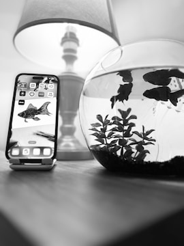 A smartphone displaying a fish illustration is placed on a stand on a wooden surface next to a fishbowl containing live fish and aquatic plants. A lamp with a fabric shade is visible in the background.