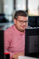 A person with glasses in a red checkered shirt is focused on working at a computer in an office setting. The environment appears professional with desks and technology visible in the background.