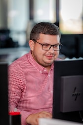 A person with glasses in a red checkered shirt is focused on working at a computer in an office setting. The environment appears professional with desks and technology visible in the background.