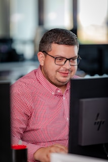 A person with glasses in a red checkered shirt is focused on working at a computer in an office setting. The environment appears professional with desks and technology visible in the background.