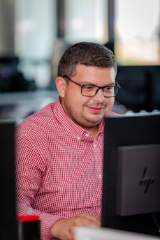 A person with glasses in a red checkered shirt is focused on working at a computer in an office setting. The environment appears professional with desks and technology visible in the background.