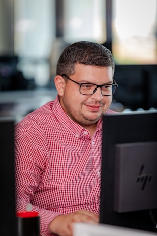 A person with glasses in a red checkered shirt is focused on working at a computer in an office setting. The environment appears professional with desks and technology visible in the background.