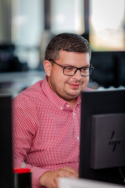 A person with glasses in a red checkered shirt is focused on working at a computer in an office setting. The environment appears professional with desks and technology visible in the background.
