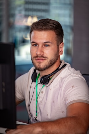 A man sits in front of a computer monitor, focused on the screen. He has short brown hair and a beard, wearing a white shirt with a pattern, and a green lanyard. Headphones hang around his neck. The background appears to be an office setting with blurred elements indicating window reflections.