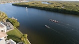 Aerial view of a wide river with two rowing boats moving along, passing by a residential area with a house and screened pool enclosure. The opposite bank is lined with dense green foliage, giving way to open waterways stretching into the distance.