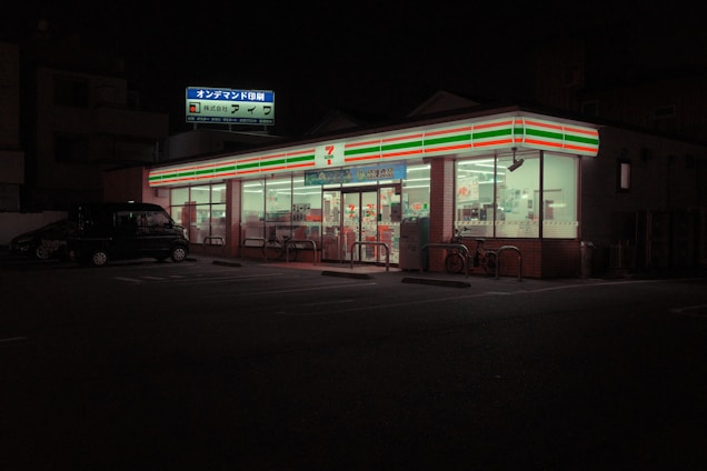 A brightly lit convenience store is visible at night, with a recognizable striped logo on its signage. The store has large glass windows and is surrounded by a darkened parking lot, with a vehicle and bicycles near the entrance. Inside, shelves and products are dimly visible under fluorescent lighting.
