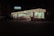 A brightly lit convenience store is visible at night, with a recognizable striped logo on its signage. The store has large glass windows and is surrounded by a darkened parking lot, with a vehicle and bicycles near the entrance. Inside, shelves and products are dimly visible under fluorescent lighting.