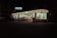 A brightly lit convenience store is visible at night, with a recognizable striped logo on its signage. The store has large glass windows and is surrounded by a darkened parking lot, with a vehicle and bicycles near the entrance. Inside, shelves and products are dimly visible under fluorescent lighting.