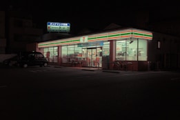 A brightly lit convenience store is visible at night, with a recognizable striped logo on its signage. The store has large glass windows and is surrounded by a darkened parking lot, with a vehicle and bicycles near the entrance. Inside, shelves and products are dimly visible under fluorescent lighting.