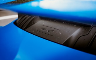 A close-up of a blue sports car's engine cover or hood. The surface is slightly dusty and wet with water droplets. The part of the car visible has a sleek, metallic design, and a number or symbol is embossed on the cover.