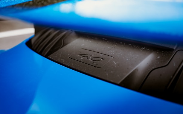 A close-up of a blue sports car's engine cover or hood. The surface is slightly dusty and wet with water droplets. The part of the car visible has a sleek, metallic design, and a number or symbol is embossed on the cover.