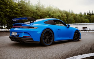 A sleek blue sports car is parked on a track, surrounded by a backdrop of lush green trees. The car features aerodynamic design elements, including a rear spoiler. The setting suggests a racing or test drive environment with a smooth asphalt surface.