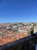 A panoramic view of a city with numerous buildings featuring red-tiled roofs spread across a hilly landscape. In the foreground, part of a stone wall and a person's hand can be seen holding a smartphone or camera. The sky is clear and blue, with sparse white clouds scattered across.