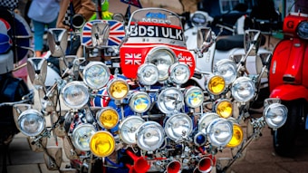 A vintage scooter adorned with an extensive array of lights and mirrors is prominently displayed. The lights vary in size, with a mix of clear and amber lenses. Several mirrors with chrome finishes are strategically positioned around the scooter. The vehicle is decorated with multiple British flags and features a license plate on the front. In the background, other scooters can be seen, some with vivid colors like red and blue.