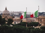 A picturesque view of a cityscape featuring a notable dome structure in the background, flanked by two tall flag poles displaying Italian flags with their distinctive green, white, and red colors. Surrounding the architectural elements are clusters of lush greenery and trees, which contrast with the warm, neutral tones of the buildings.