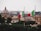 A picturesque view of a cityscape featuring a notable dome structure in the background, flanked by two tall flag poles displaying Italian flags with their distinctive green, white, and red colors. Surrounding the architectural elements are clusters of lush greenery and trees, which contrast with the warm, neutral tones of the buildings.