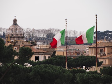 A picturesque view of a cityscape featuring a notable dome structure in the background, flanked by two tall flag poles displaying Italian flags with their distinctive green, white, and red colors. Surrounding the architectural elements are clusters of lush greenery and trees, which contrast with the warm, neutral tones of the buildings.