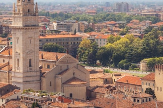 An aerial view of an urban landscape featuring a prominent historical tower with intricate architectural details. Surrounding buildings are characterized by red-tiled roofs. Lush greenery and trees are scattered throughout the area, with modern apartment buildings visible in the background.