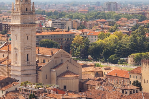 An aerial view of an urban landscape featuring a prominent historical tower with intricate architectural details. Surrounding buildings are characterized by red-tiled roofs. Lush greenery and trees are scattered throughout the area, with modern apartment buildings visible in the background.