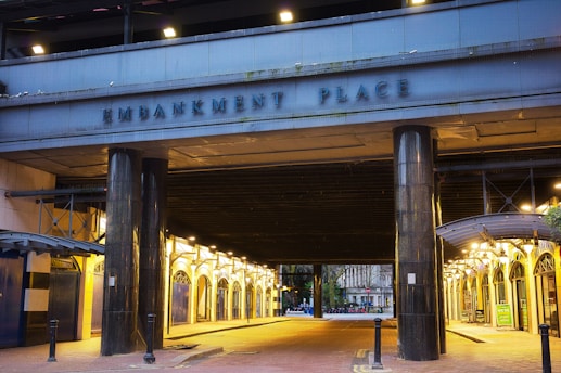 A large, illuminated archway labeled 'Embankment Place' leads into a covered walkway or entrance. The structure features dark pillars and is flanked by rows of light fixtures, casting a warm glow on the pavement. In the background, trees and a distant building are visible.