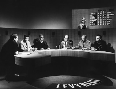 A group of six individuals seated around a curved table, engaged in discussion. The setting appears to be a television studio with a large, semicircular desk. Behind the group, a large screen shows a list of names and numbers, along with a woman standing. The overall atmosphere suggests a formal or televised panel discussion or game show.