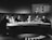 A group of six individuals seated around a curved table, engaged in discussion. The setting appears to be a television studio with a large, semicircular desk. Behind the group, a large screen shows a list of names and numbers, along with a woman standing. The overall atmosphere suggests a formal or televised panel discussion or game show.