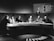 A group of six individuals seated around a curved table, engaged in discussion. The setting appears to be a television studio with a large, semicircular desk. Behind the group, a large screen shows a list of names and numbers, along with a woman standing. The overall atmosphere suggests a formal or televised panel discussion or game show.