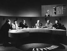 A group of six individuals seated around a curved table, engaged in discussion. The setting appears to be a television studio with a large, semicircular desk. Behind the group, a large screen shows a list of names and numbers, along with a woman standing. The overall atmosphere suggests a formal or televised panel discussion or game show.
