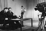 Two men are sitting at a table, engaged in conversation or possibly conducting an interview. A large television camera is positioned towards them, suggesting they are being filmed for a broadcast. The room is furnished with curtains in the background, and a vase of flowers is placed on the table.