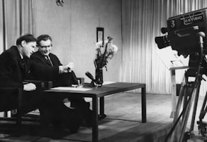 Two men are sitting at a table, engaged in conversation or possibly conducting an interview. A large television camera is positioned towards them, suggesting they are being filmed for a broadcast. The room is furnished with curtains in the background, and a vase of flowers is placed on the table.