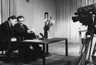 Two men are sitting at a table, engaged in conversation or possibly conducting an interview. A large television camera is positioned towards them, suggesting they are being filmed for a broadcast. The room is furnished with curtains in the background, and a vase of flowers is placed on the table.