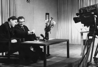 Two men are sitting at a table, engaged in conversation or possibly conducting an interview. A large television camera is positioned towards them, suggesting they are being filmed for a broadcast. The room is furnished with curtains in the background, and a vase of flowers is placed on the table.