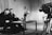 Two men are sitting at a table, engaged in conversation or possibly conducting an interview. A large television camera is positioned towards them, suggesting they are being filmed for a broadcast. The room is furnished with curtains in the background, and a vase of flowers is placed on the table.