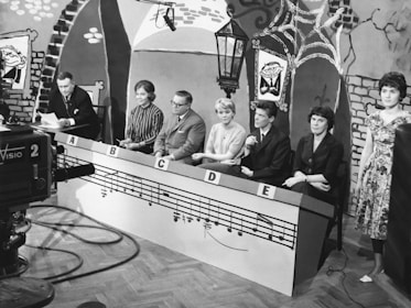 A black and white television studio scene with seven individuals seated behind a panel labeled with letters A to E. The set is decorated with playful drawings, including a large cartoonish face, bricks, and musical notes. A large camera is positioned in the foreground, aimed at the panelists. A vintage microphone hangs from above, and the environment has an old-fashioned aesthetic.
