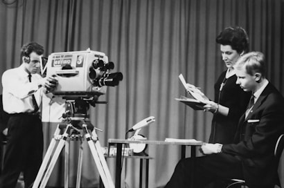 A black and white photo of a television studio, featuring a cameraman adjusting a large camera labeled 'Es Viso' with the number '3'. Beside him, a seated young man wearing a suit is listening intently to a woman who stands next to him, holding a script or paper. The scene is set indoors, likely around the mid-20th century, with a curtain as the backdrop.