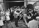 A vintage television studio setting with a large group of people gathered around a cameraman operating an old-fashioned television camera. The audience is dressed in formal attire typical of the mid-20th century. The studio has a brick wall backdrop with cartoonish artwork.