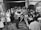 A vintage television studio setting with a large group of people gathered around a cameraman operating an old-fashioned television camera. The audience is dressed in formal attire typical of the mid-20th century. The studio has a brick wall backdrop with cartoonish artwork.
